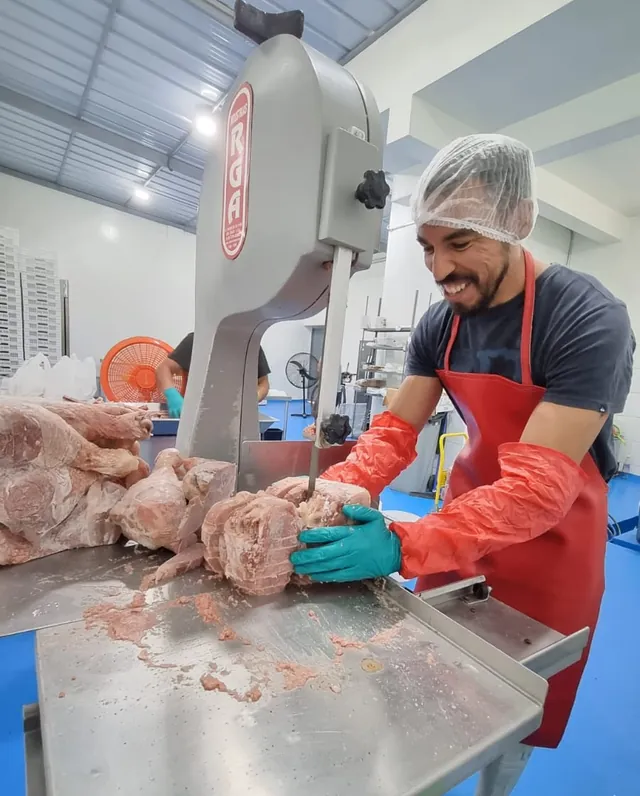 Trabajador con cofia y delantal rojo cortando carne y hueso en una sierra de cinta.