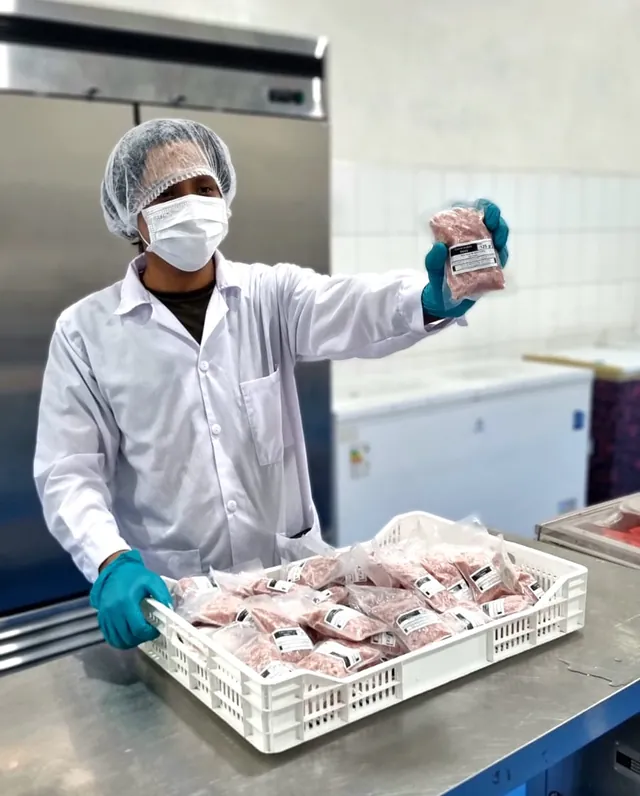 Trabajador con mascarilla mostrando una bolsa de comida empacada, con una caja llena de bolsas delante.