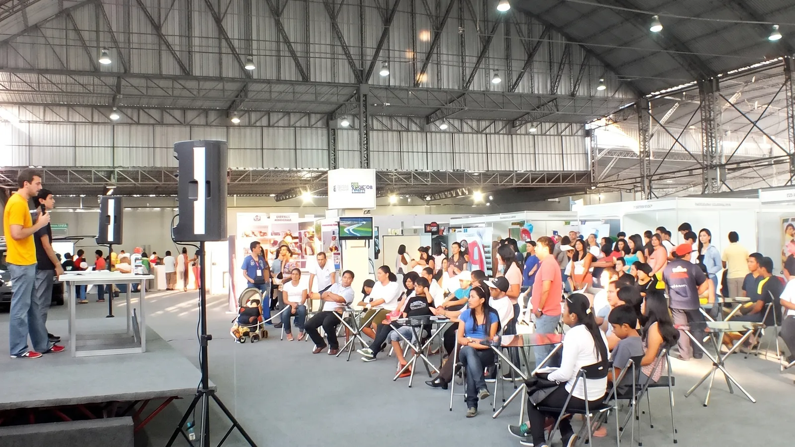 Público sentado escuchando una charla en una feria, dentro de un gran pabellón con stands.